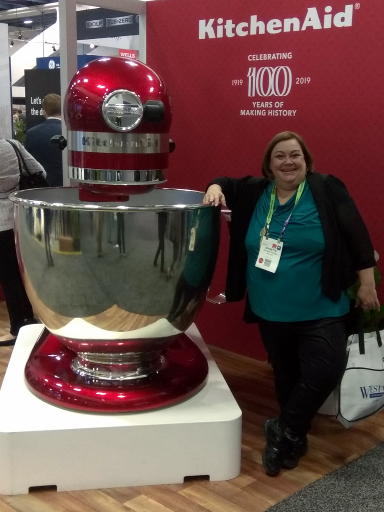 Jeanette stands next to a giant KitchenAid mixer at a trade show.